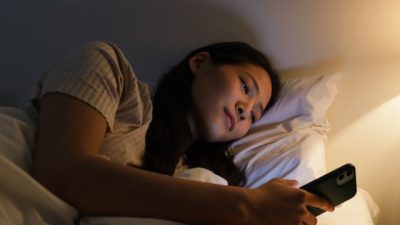 Seorang wanita berbaring di tempat tidur di malam hari menggunakan smartphone / pexels-ron-lach